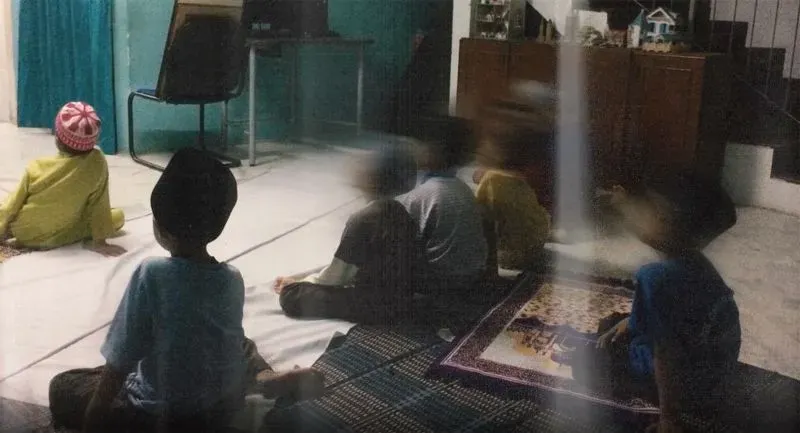 مجموعة من الأطفال يجلسون في غرفة مظلمة يشاهدون التلفاز، مع ظهور بعض تفاصيل الغرفة مثل السجاد والأثاث، مما يعكس بيئة غير آمنة.