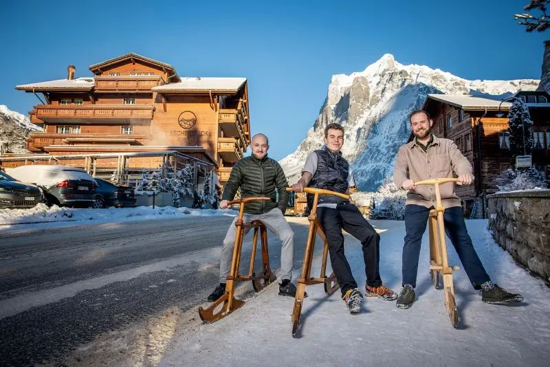 ثلاثة رجال يجلسون على زلاجات خشبية تقليدية في منطقة ثلجية، مع خلفية جبال الألب ومباني خشبية في قرية سويسرية.