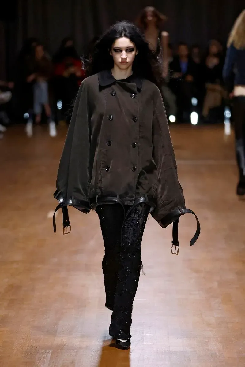 عارضة أزياء تسير على منصة عرض، ترتدي سترة فضفاضة بلون بني داكن مع تفاصيل جلدية، بينما تتجه الأنظار نحو تصميمها الفريد والمبتكر.