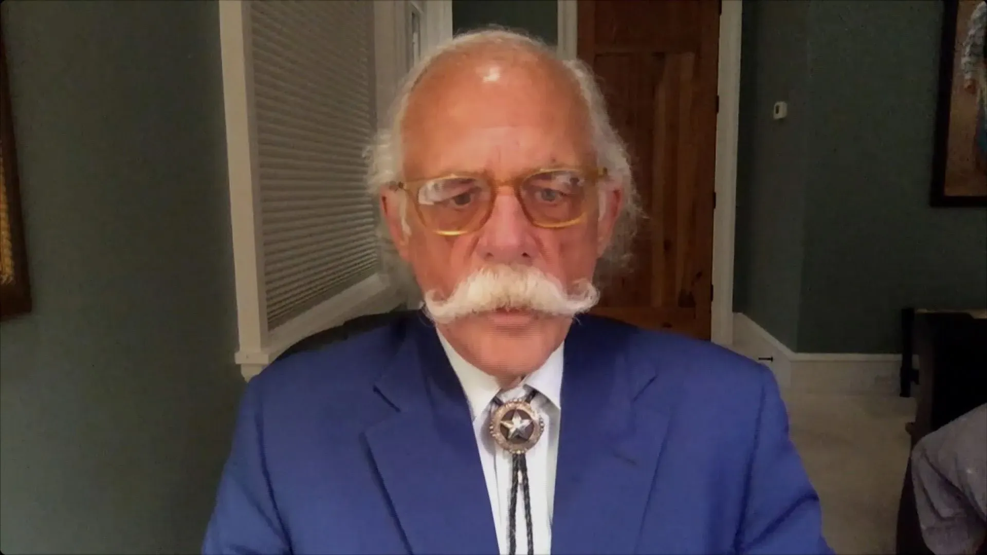 رجل مسن ذو شارب كبير يرتدي بدلة زرقاء ونظارات، يجلس أمام خلفية محايدة، يتحدث في جلسة عبر الفيديو.