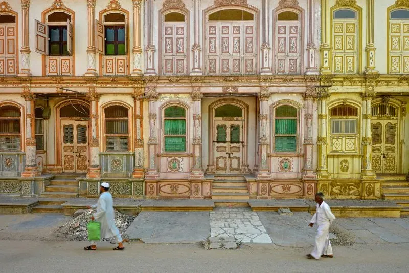 واجهة تاريخية لقصور سيدهبور، تظهر تفاصيل معمارية فريدة وألوان باهتة، مع مرور شخصين في الشارع.