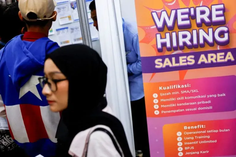 شابة ترتدي حجابًا تسير بجوار لوحة إعلانات تعلن عن وظائف شاغرة في منطقة المبيعات، تعكس تحديات البطالة بين الشباب في إندونيسيا.