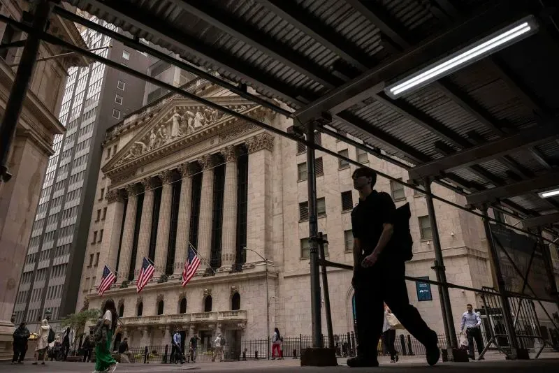 صورة لمبنى بورصة نيويورك مع علم الولايات المتحدة، حيث يسير الناس في الشارع. تعكس الصورة نشاط السوق المالي وتأثير التعريفات الجمركية.