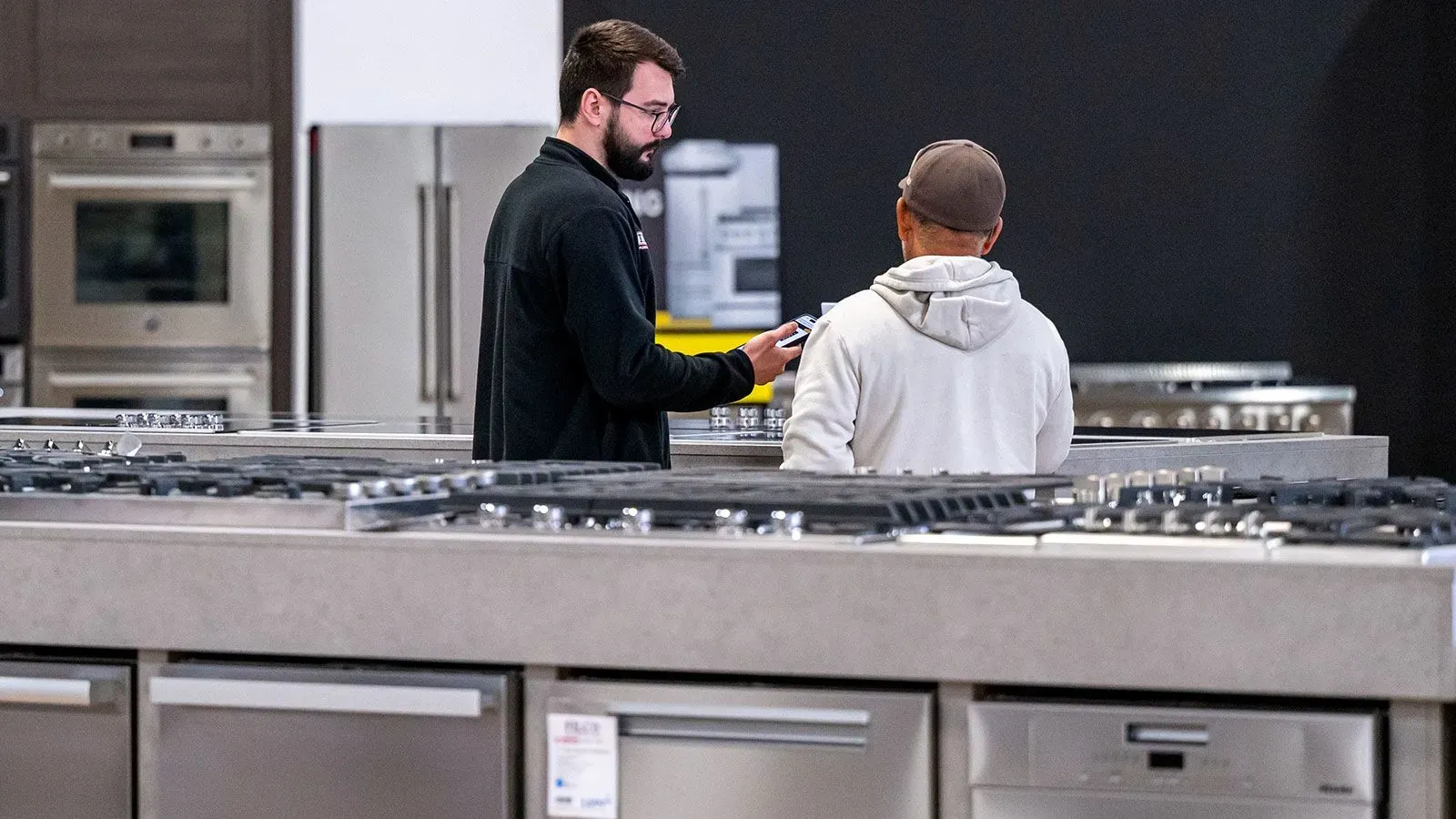 شخصان يتحدثان في متجر للأجهزة المنزلية، مع وجود معدات مطبخ حديثة في الخلفية، مما يعكس اتجاهات السوق الحالية.