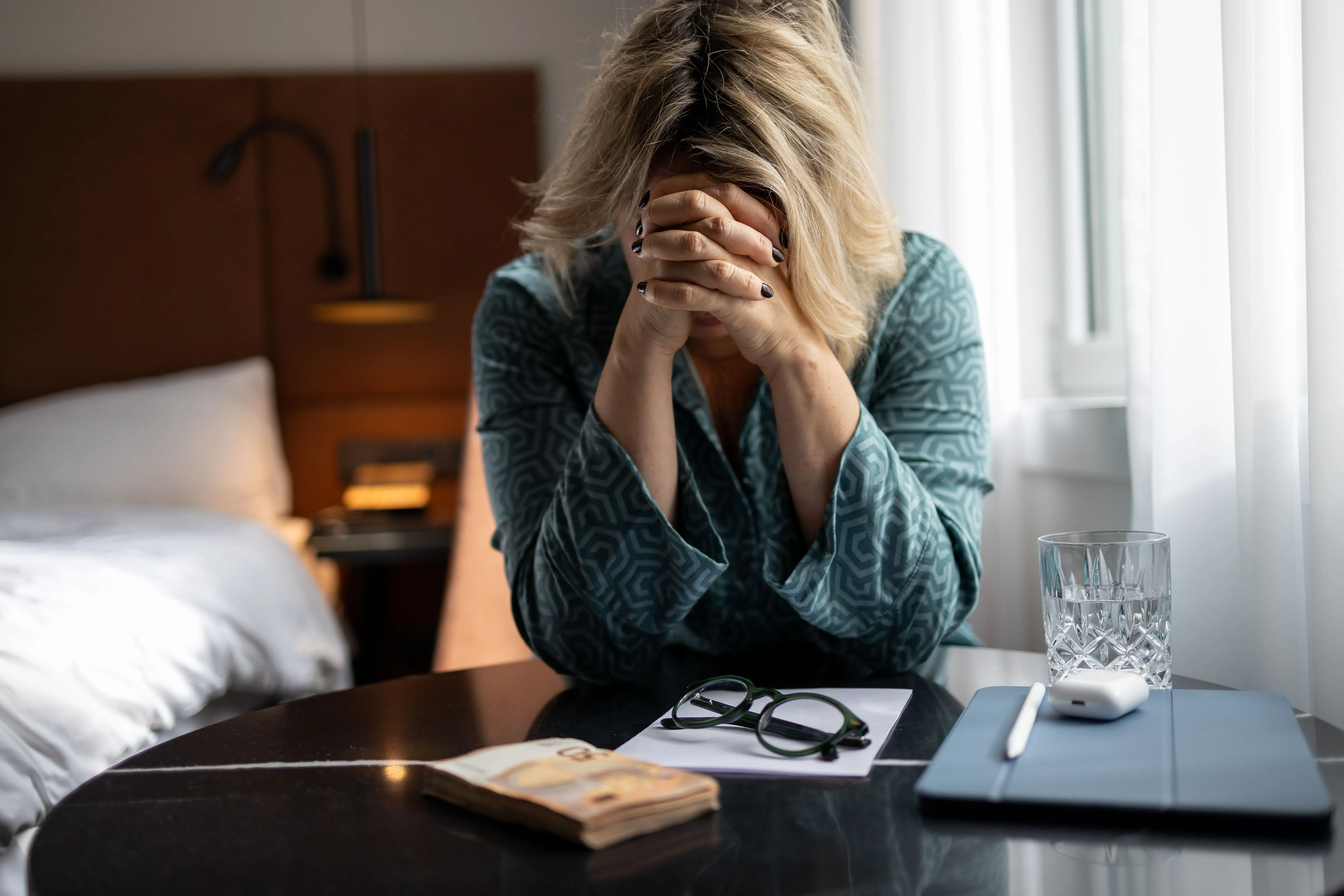 امرأة تشعر بالتوتر، تدعم رأسها بيدها، وتجلس أمام طاولة تحتوي على نظارات وكوب ماء، في بيئة منزلية مريحة.