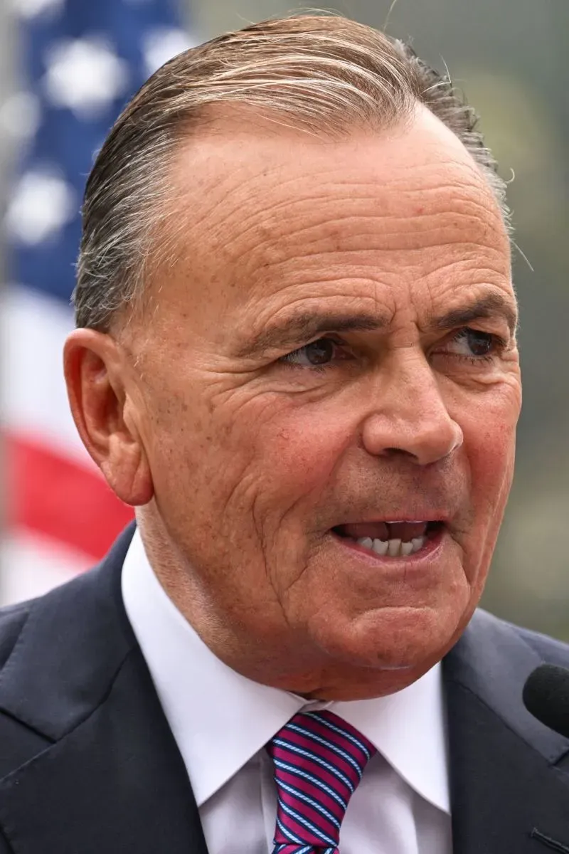 صورة لرجل يتحدث بجدية، يرتدي بدلة داكنة مع ربطة عنق ملونة، ويظهر خلفه علم أمريكي. تعكس تعابير وجهه القلق بشأن القضايا الاقتصادية.