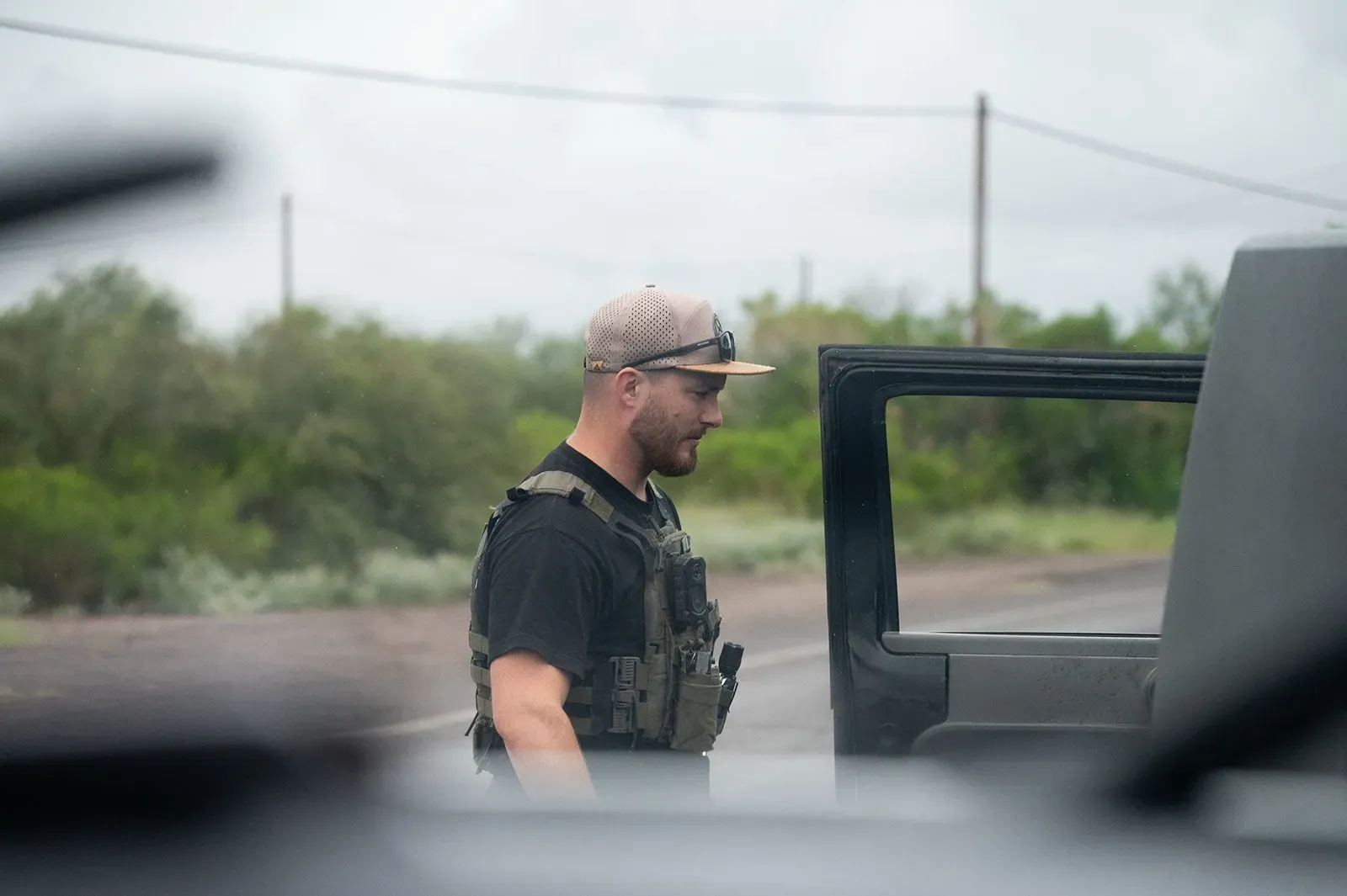 رجل يرتدي سترة واقية وقبعة، يقف بجانب سيارة في منطقة صحراوية، في سياق تحقيق حول تهريب المهاجرين.