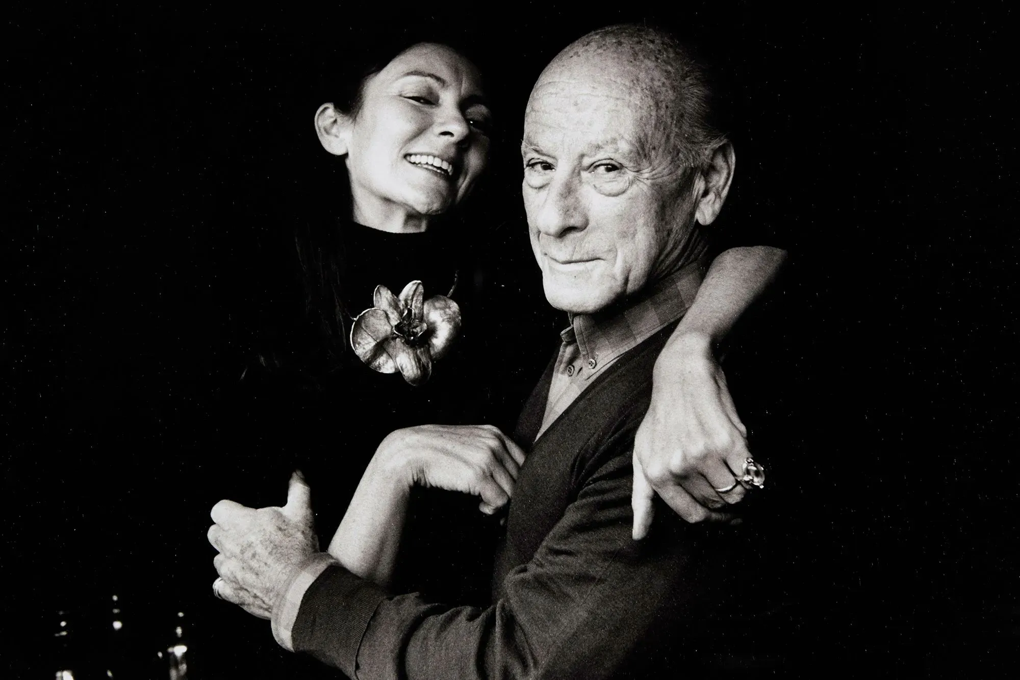 صورة بالأبيض والأسود لامرأة مبتسمة تحتضن رجلًا مسنًا، مع خلفية داكنة، تعكس العلاقة الحميمية بينهما في عالم الفن.