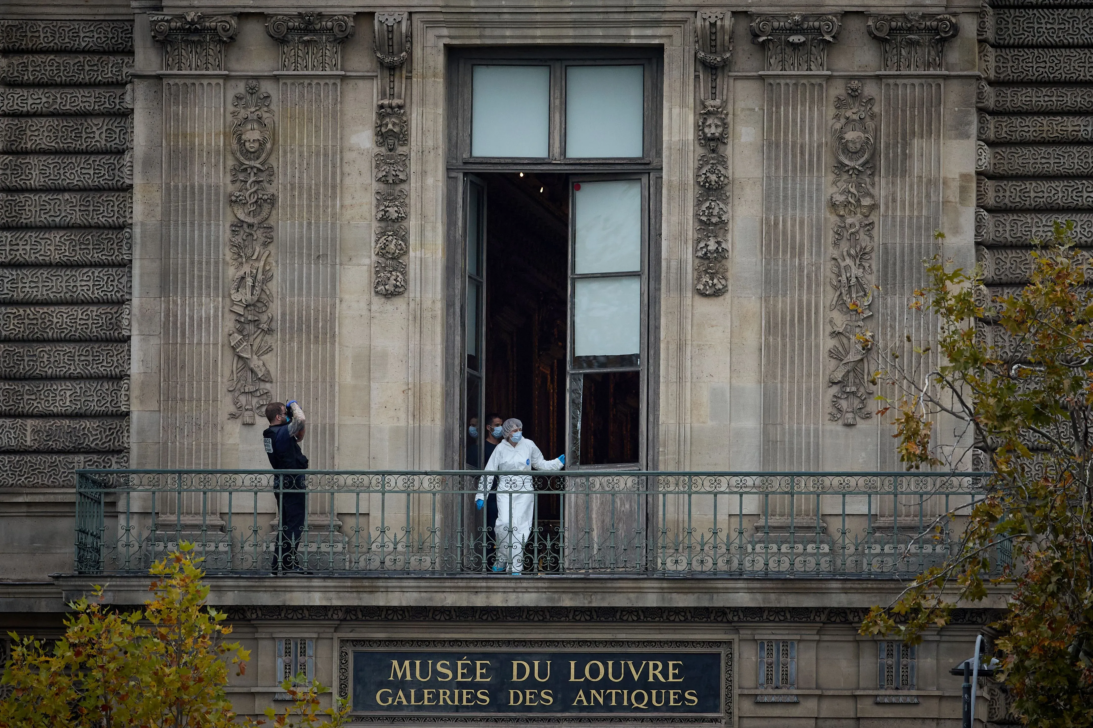 تحقيقات في سرقة متحف اللوفر، حيث يظهر المحققون في ملابس واقية أمام نافذة المتحف، مع لافتة "متحف اللوفر" واضحة.
