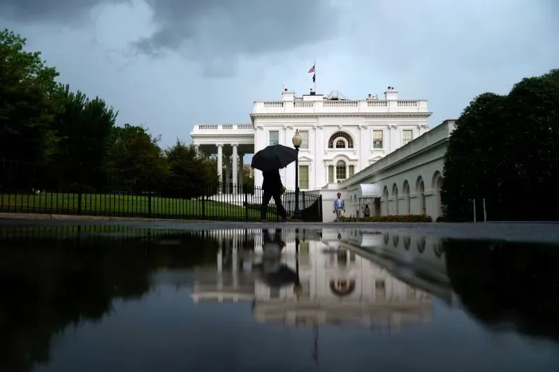رجل يحمل مظلة يسير بالقرب من البيت الأبيض تحت سماء ملبدة بالغيوم، مع انعكاس المبنى على بركة ماء.