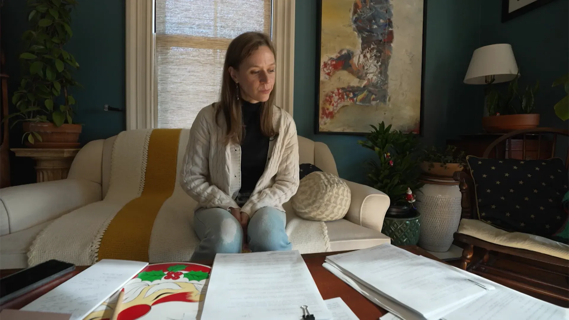 امرأة تجلس في غرفة معيشة، محاطة بأوراق ومستندات، تعبر عن القلق والتفكير في معركة للحصول على مزايا المحاربين القدامى بعد انتحار زوجها.