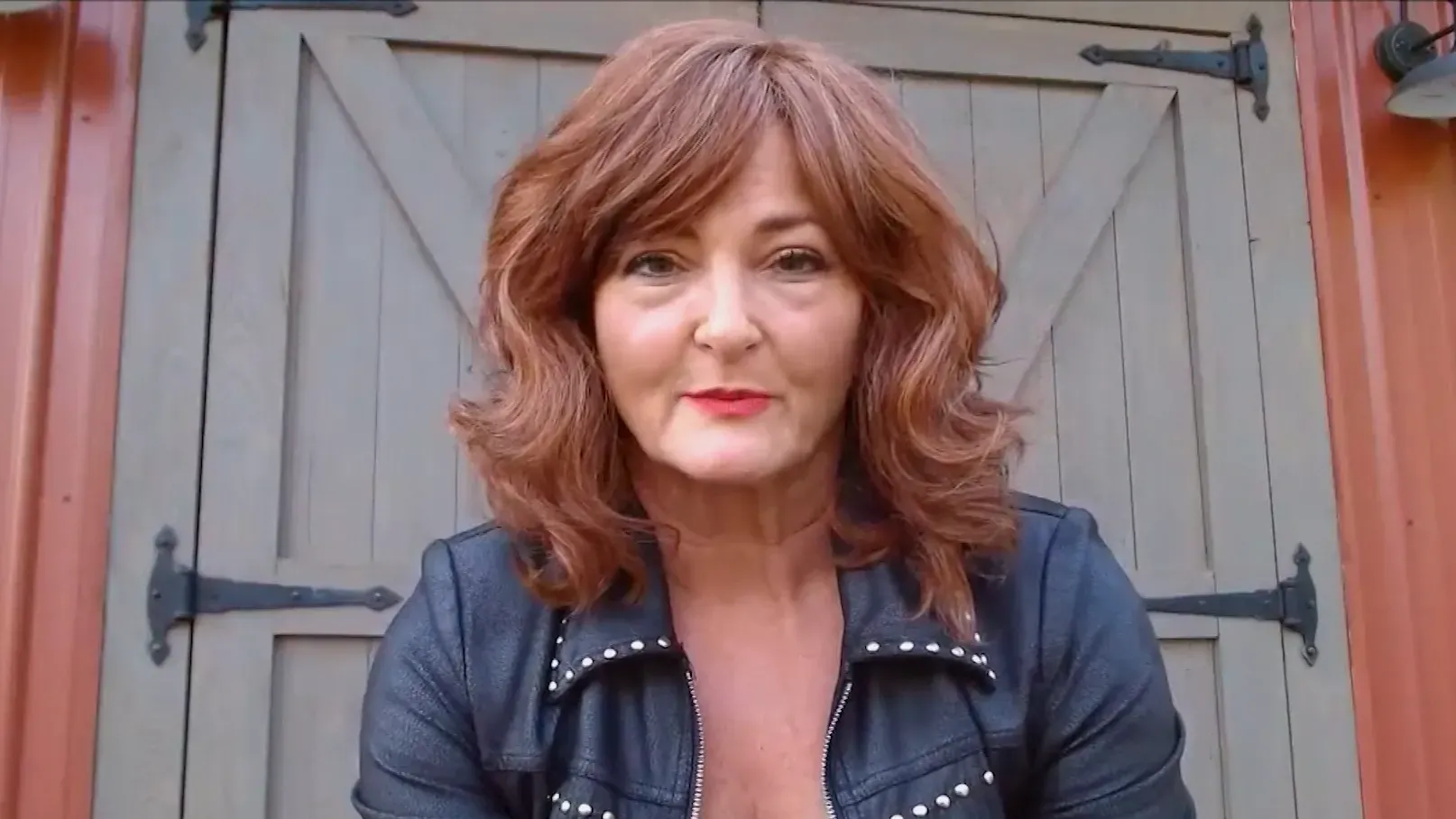 امرأة ذات شعر بني مموج وحواجب مرتفعة، ترتدي سترة جلدية، تظهر في المقدمة مع باب خشبي خلفها.