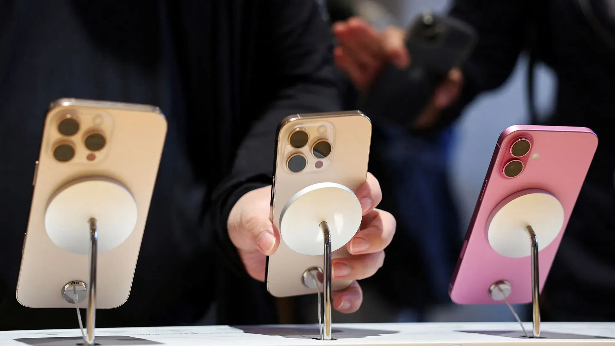 هواتف آيفون معروضة في متجر، حيث يظهر شخص يحمل أحد الهواتف. تتعلق المقالة بارتفاع أسعار الآيفون نتيجة التعريفات الجمركية.