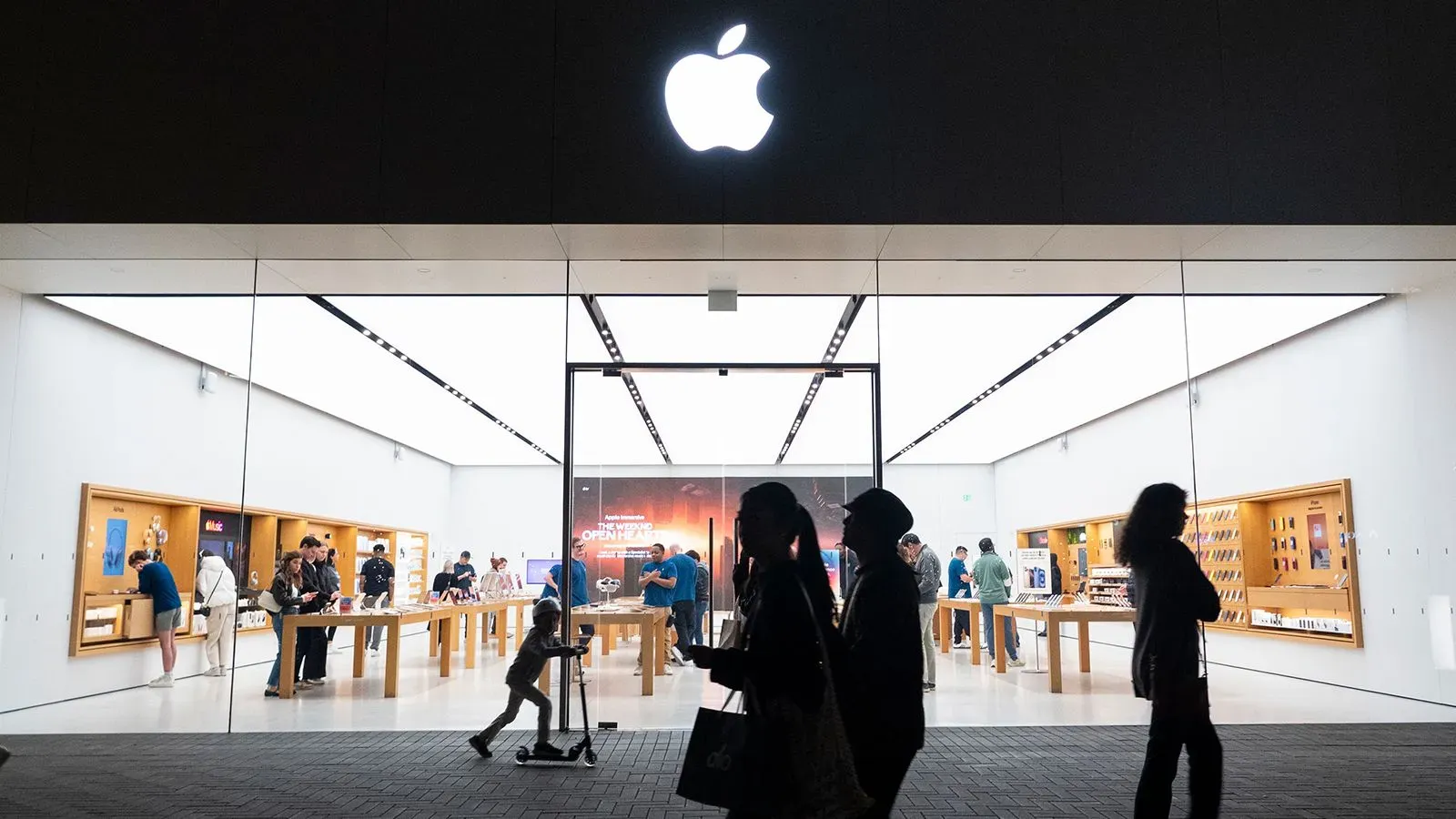 واجهة متجر آبل تعرض شعار الشركة مع زبائن يتجولون داخل المتجر، في سياق الحديث عن تأثير التعريفات الجمركية على أسعار الآيفون.