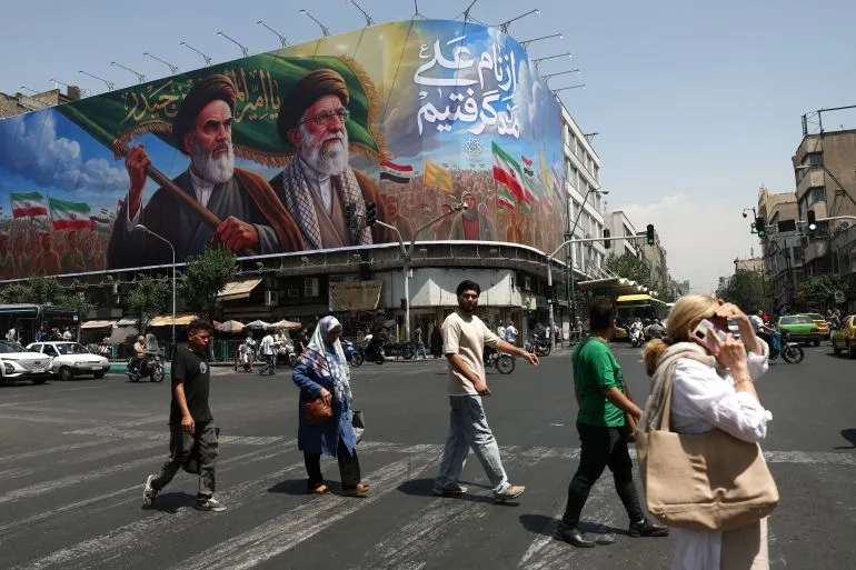 لوحة جدارية كبيرة في طهران تظهر شخصيات تاريخية إيرانية، مع أشخاص يعبرون الشارع. تعكس الصورة التوترات السياسية الحالية وتاريخ إيران.