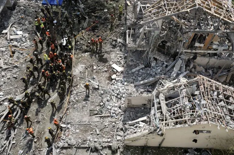 فرق الإنقاذ تعمل في موقع دمار كبير بعد الهجمات الصاروخية في إسرائيل، مع وجود أنقاض ومباني مدمرة في الخلفية.