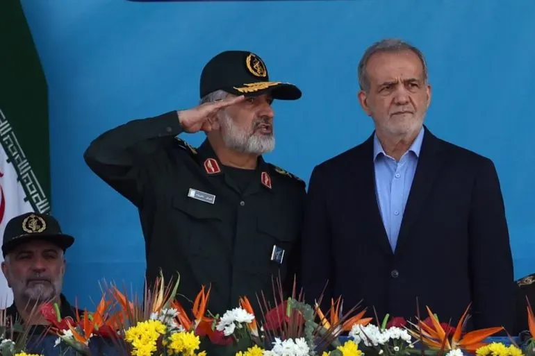 جنرال إيراني يرتدي زيًا عسكريًا ويؤدي التحية، يقف بجانب مسؤول حكومي في حدث رسمي، مع خلفية زرقاء وزهور ملونة.