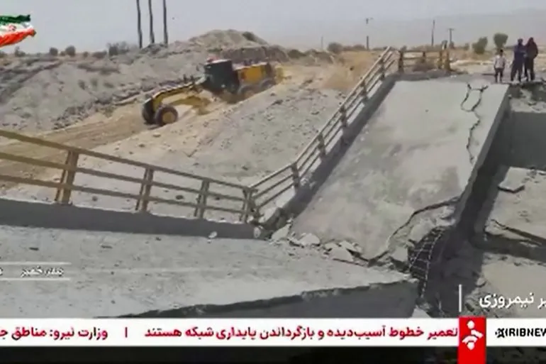 جسر مدمر في منطقة صحراوية مع وجود آليات ثقيلة وأشخاص في الخلفية، يعكس تأثير الضربات الأمريكية على البنية التحتية في إيران.