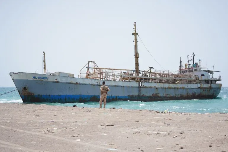 سفينة قديمة متوقفة على شاطئ باب المندب، مع وجود شخص يقف بالقرب منها، مما يبرز أهمية هذا الممر المائي الحيوي لتجارة النفط العالمية.