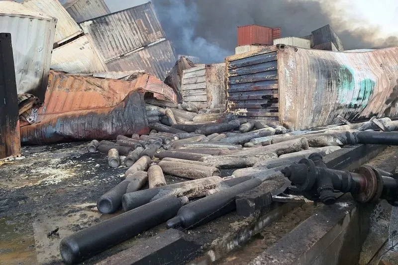 حطام حاويات مدمرة في ميناء بندر عباس بعد انفجار ضخم، مع وجود دخان كثيف في الخلفية، يشير إلى وقوع حادث مأسوي مرتبط بالمواد الكيميائية.