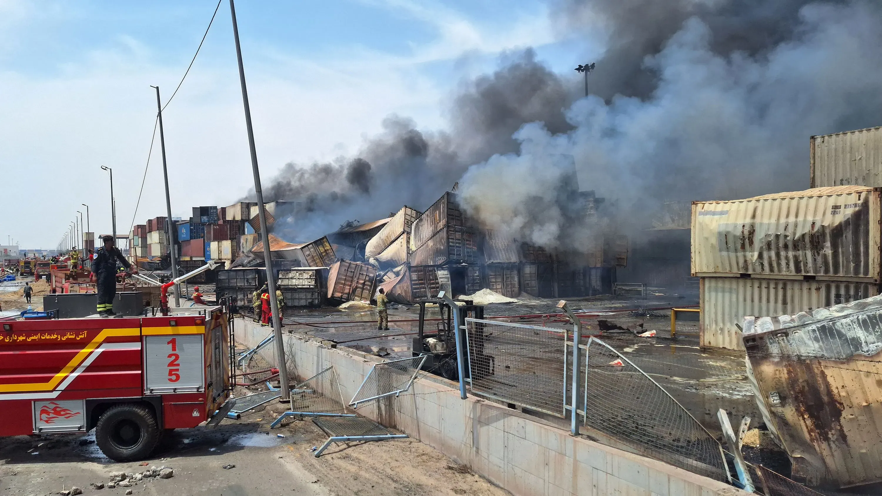 دخان كثيف يتصاعد من حاويات شحن متضررة في ميناء بندر عباس بعد انفجار ضخم، مع وجود رجال إطفاء في الموقع.