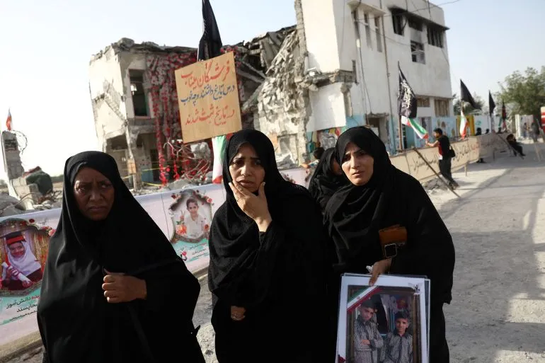 نساء يرتدين الحجاب يقفن أمام مبانٍ مدمّرة، يحملن صورًا لأحبائهن، في سياق الاحتجاجات على الأوضاع الإنسانية في المنطقة.