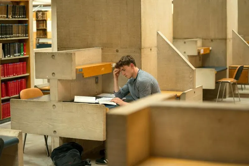 طالب يدرس في مكتبة كلية ترينيتي دبلن، محاط بمقاعد دراسية مصممة من الخرسانة، تعكس العمارة الوحشية للمبنى.
