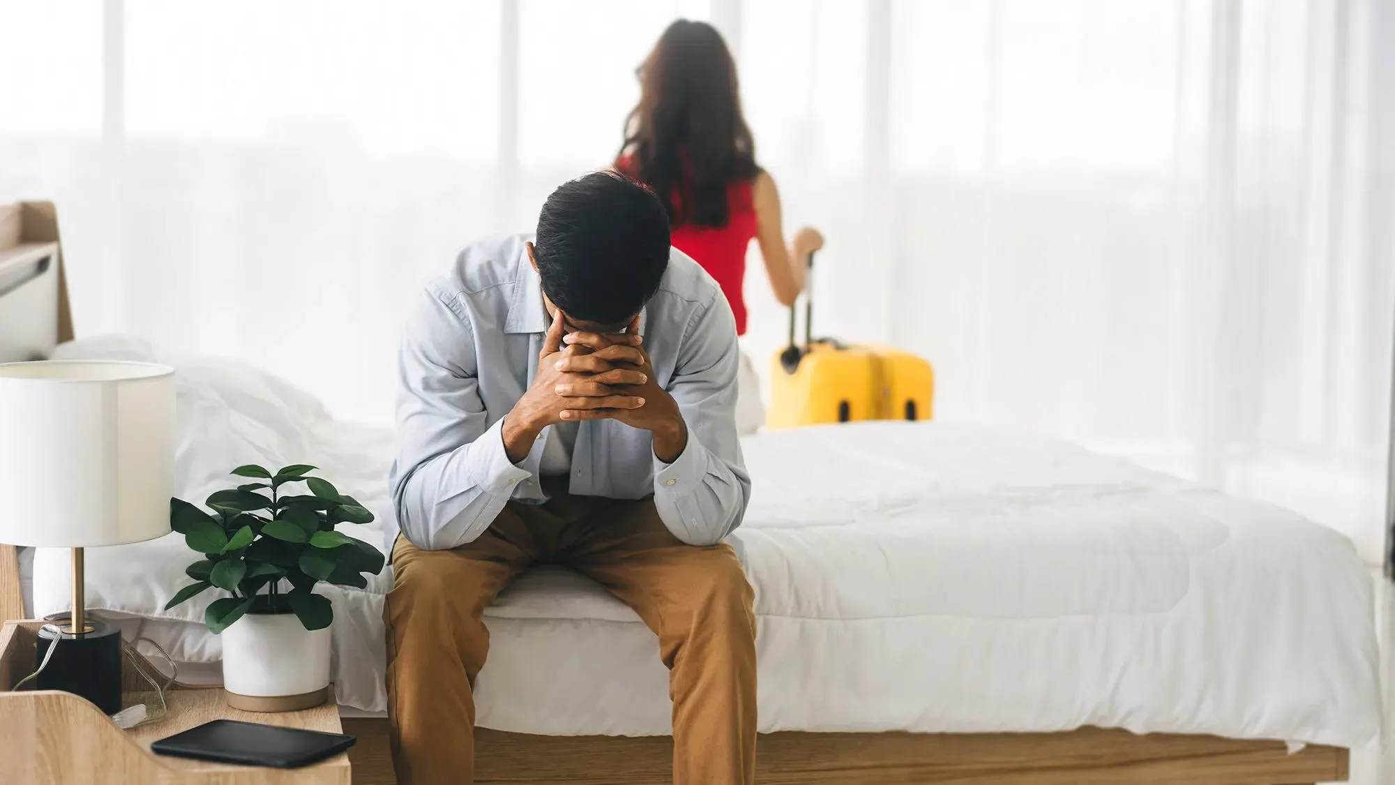 رجل يجلس حزينًا على حافة السرير بينما تتركه امرأة تحمل حقيبة سفر، مما يعكس مشاعر الانفصال أو الطلاق.