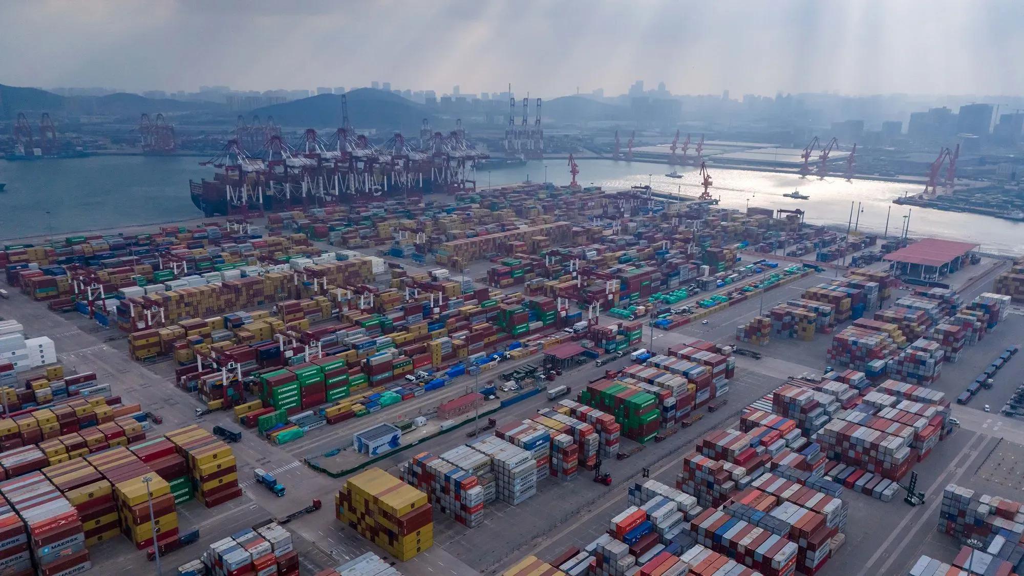 صورة لميناء شحن مزدحم يحتوي على حاويات ملونة، مع سفن وشاحنات، تعكس تأثير الرسوم الجمركية على التجارة الأمريكية.