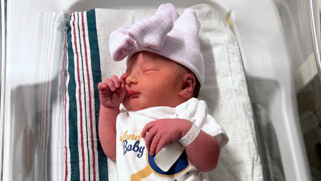 طفل حديث الولادة يرتدي قميصًا يحمل عبارة "طفل الجزيرة" مع قبعة كبيرة، مستلقيًا في سرير مستشفى.