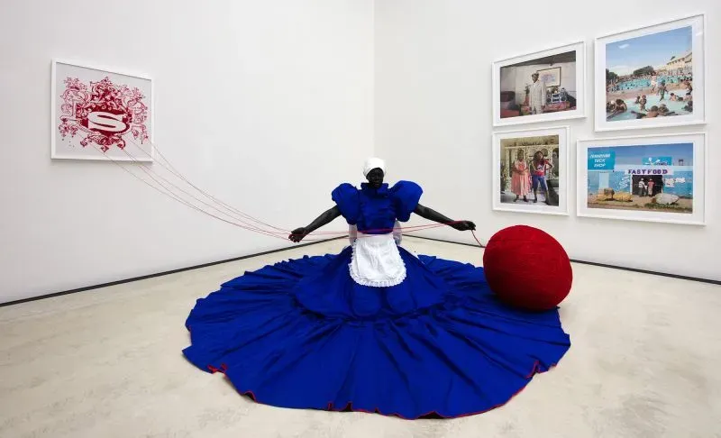 عرض فني لشخصية ترتدي فستانًا أزرق كبيرًا مع مئزر أبيض، محاطة بصور تعكس الثقافة والتاريخ، تعبر عن الهوية والطبقة.