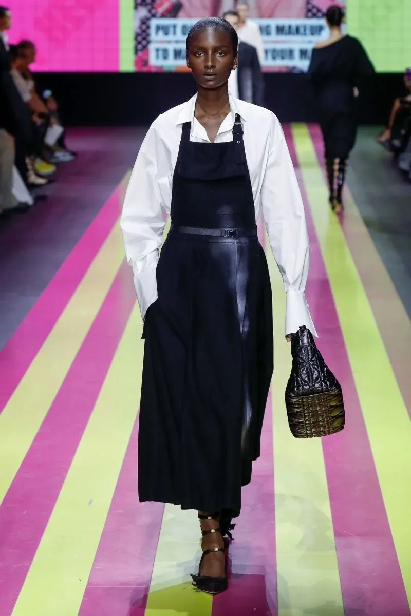 عارضة أزياء ترتدي مئزرًا أسود مع قميص أبيض، تمشي على ممر عرض ملون، مما يعكس تأثير المئزر في الثقافة الشعبية والموضة الحديثة.