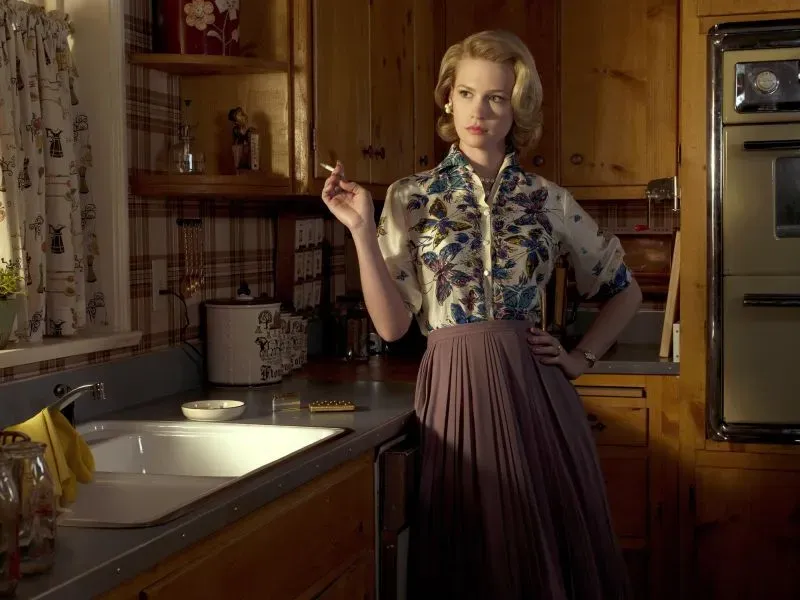 امرأة ترتدي قميصًا مزخرفًا وتنورة طويلة في مطبخ خشبي، تدخن سيجارة، تعكس أجواء الحياة المنزلية التقليدية.