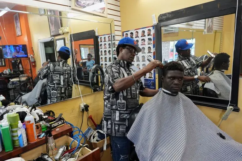 حلاق يعمل على تسريح شعر زبون في صالون حلاقة، مع وجود صور لقصات شعر على الحائط وأدوات حلاقة متنوعة على الطاولة.