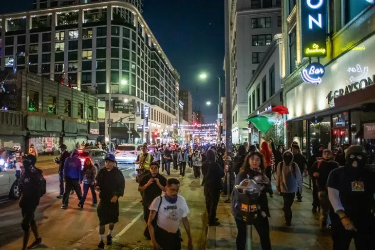 احتجاجات في لوس أنجلوس ليلاً، حيث يسير المتظاهرون في الشارع، مع وجود علم مكسيكي يرفرف في الخلفية، وسط أضواء المدينة.