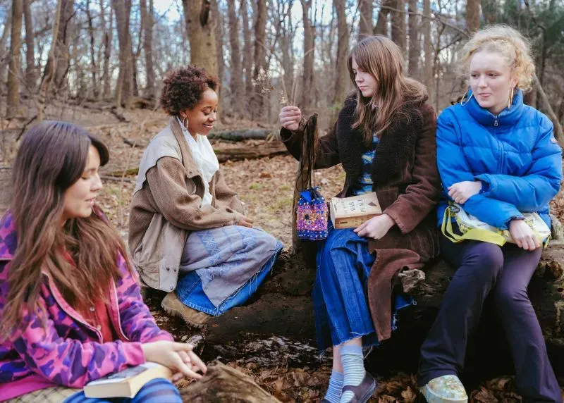 مجموعة من أربع نساء يجلسن في غابة، يتبادلن الحديث حول الكتب والتواصل، مع أجواء طبيعية هادئة تعكس تأثير وسائل التواصل الاجتماعي.