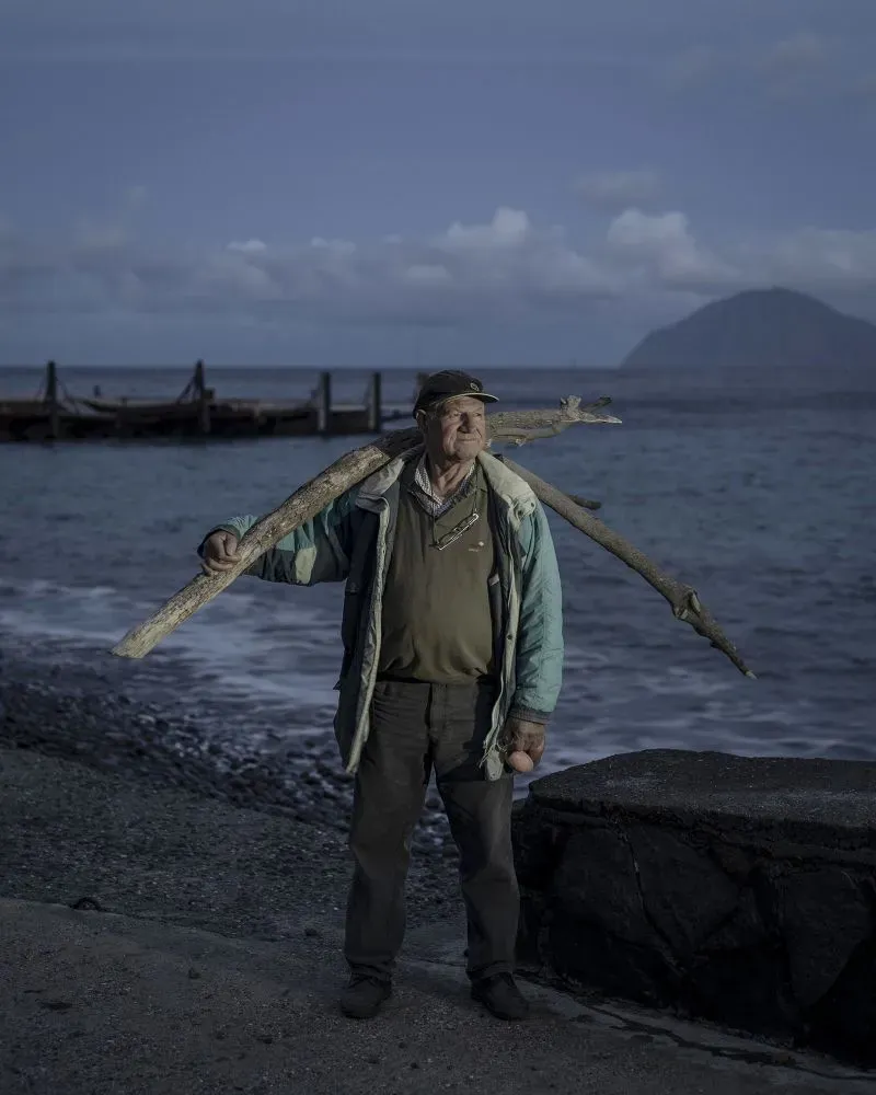 رجل يحمل عصا كبيرة على كتفه، واقفاً على شاطئ جزيرة أليكودي الهادئة، مع مناظر طبيعية بركانية جيئة وذهاباً في الخلفية.