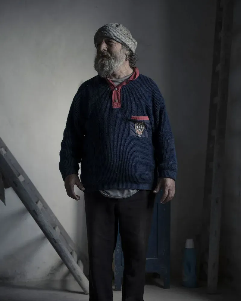 رجل مسن يرتدي سترة صوفية وقبعة، يقف في مكان مغلق مع إضاءة خافتة، مما يعكس الحياة التقليدية في جزيرة أليكودي النائية.
