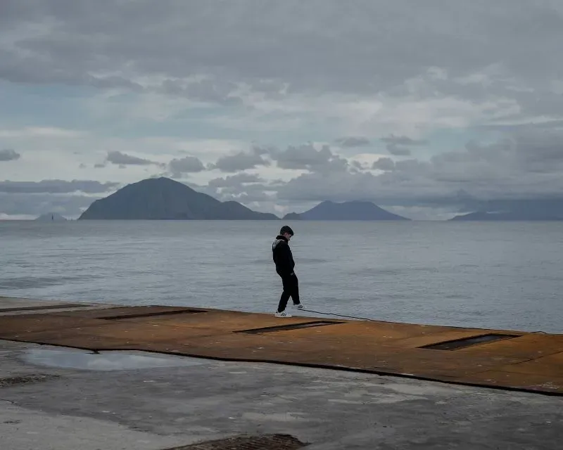 رجل يسير على رصيف في جزيرة أليكودي الإيطالية، تظهر في الخلفية جبال وسماء غائمة، تعكس شعور العزلة والهدوء.