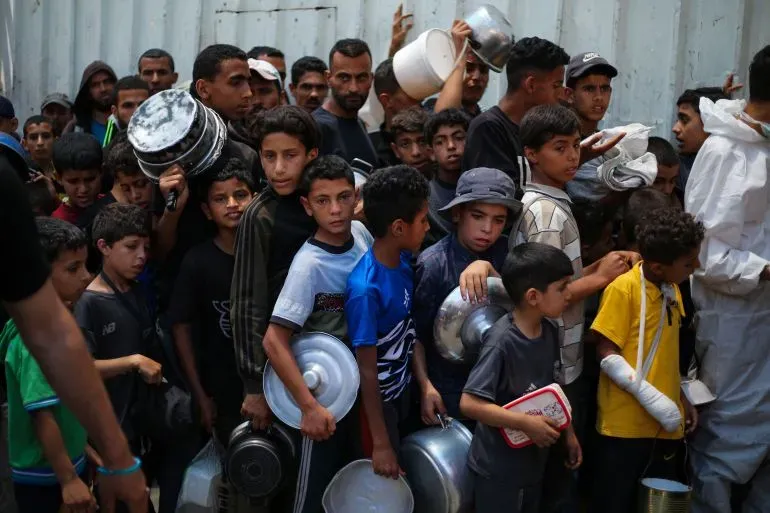 أطفال فلسطينيون يتجمعون في طابور للحصول على الطعام، مع وجود بالغين في الخلفية، في سياق الأزمة الإنسانية المستمرة في غزة.