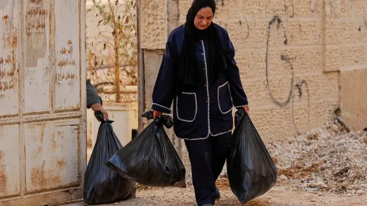 امرأة فلسطينية تحمل أكياس قمامة في منطقة تضررت من النزاع، تعكس الأثر الإنساني للأحداث الجارية في الضفة الغربية.