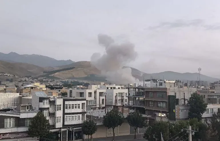 دخان يتصاعد من موقع انفجار في منطقة جبلية بالقرب من مدينة إيرانية، مع مشاهد للدمار في المباني السكنية المحيطة.