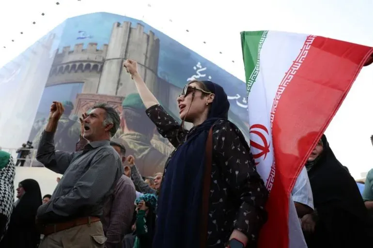 محتجون إيرانيون يرفعون الأعلام ويهتفون في تجمع حاشد، مع خلفية تضم لوحة جدارية كبيرة تعكس التوترات السياسية.