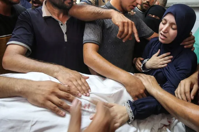 مشهد مأساوي لعائلة تجمع حول جثمان أحد أفرادها، حيث تظهر مشاعر الحزن والألم في الوجوه والأيدي الممدودة.