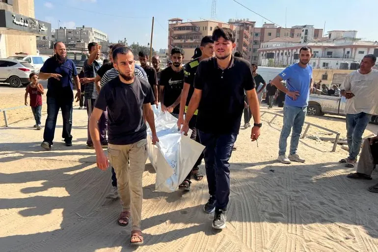 رجال يحملون جثمانًا مغطى في غزة وسط حشد من الناس، مع خلفية لمبانٍ متضررة، تعبيرًا عن الأثر المدمر للهجمات الإسرائيلية.