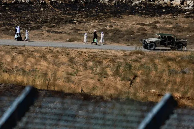 مجموعة من الأشخاص يسيرون على طريق بالقرب من مركبة عسكرية، مع خلفية من الأراضي المحترقة، في سياق التوترات العسكرية في المنطقة.