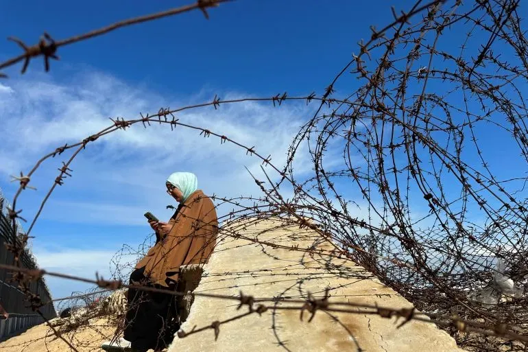 امرأة ترتدي حجابًا، تجلس على جدار محاط بسياج شائك، وتستخدم هاتفها الذكي، مع سماء زرقاء في الخلفية.