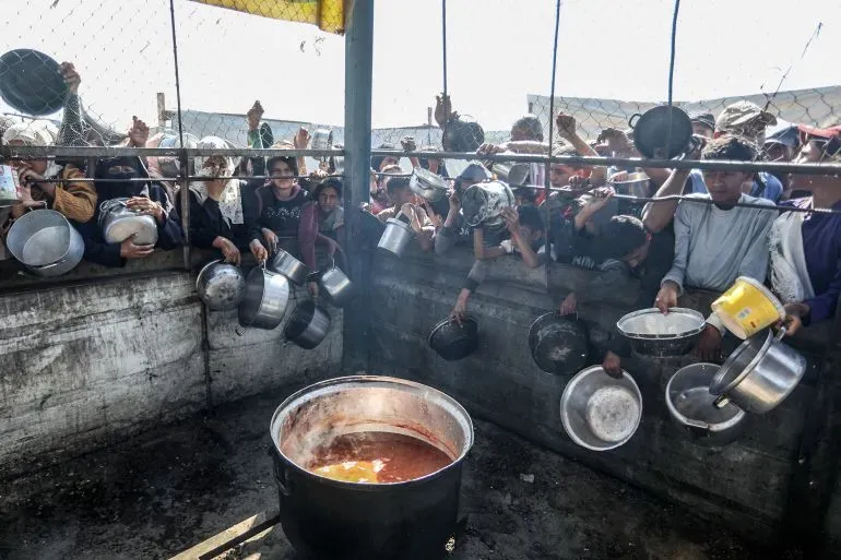 مشهد لجماهير من الفلسطينيين في غزة يتجمعون حول قدر كبير للطعام، حاملين أوعية فارغة، في سياق أزمة إنسانية متفاقمة.