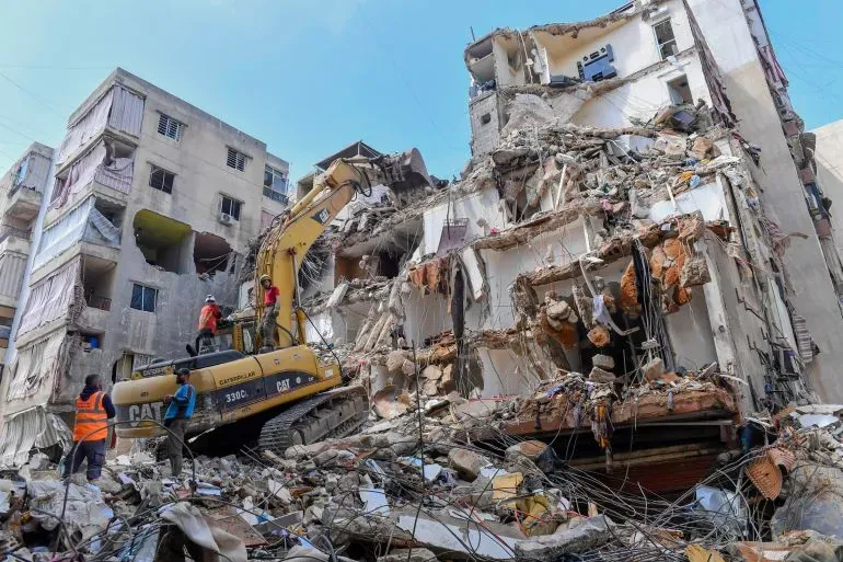 تظهر الصورة عمارة مدمّرة في لبنان، حيث يعمل عمال الإنقاذ باستخدام جرافة لإزالة الأنقاض بعد الغارات الإسرائيلية، مما يعكس تأثير الصراع المستمر.