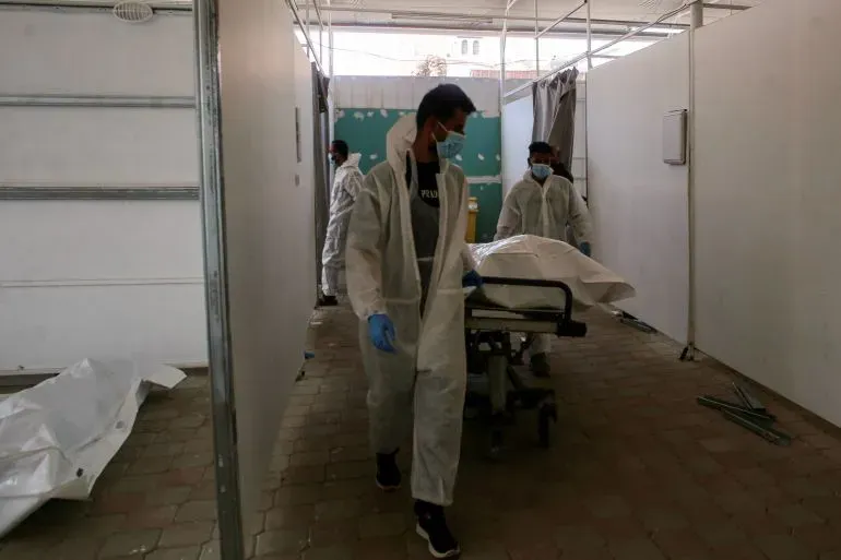 عاملون في مجال الطب يحملون جثة فلسطيني في مرفق طبي، وسط آثار تعذيب على بعض الجثث، في سياق إعادة الجثامين إلى غزة.